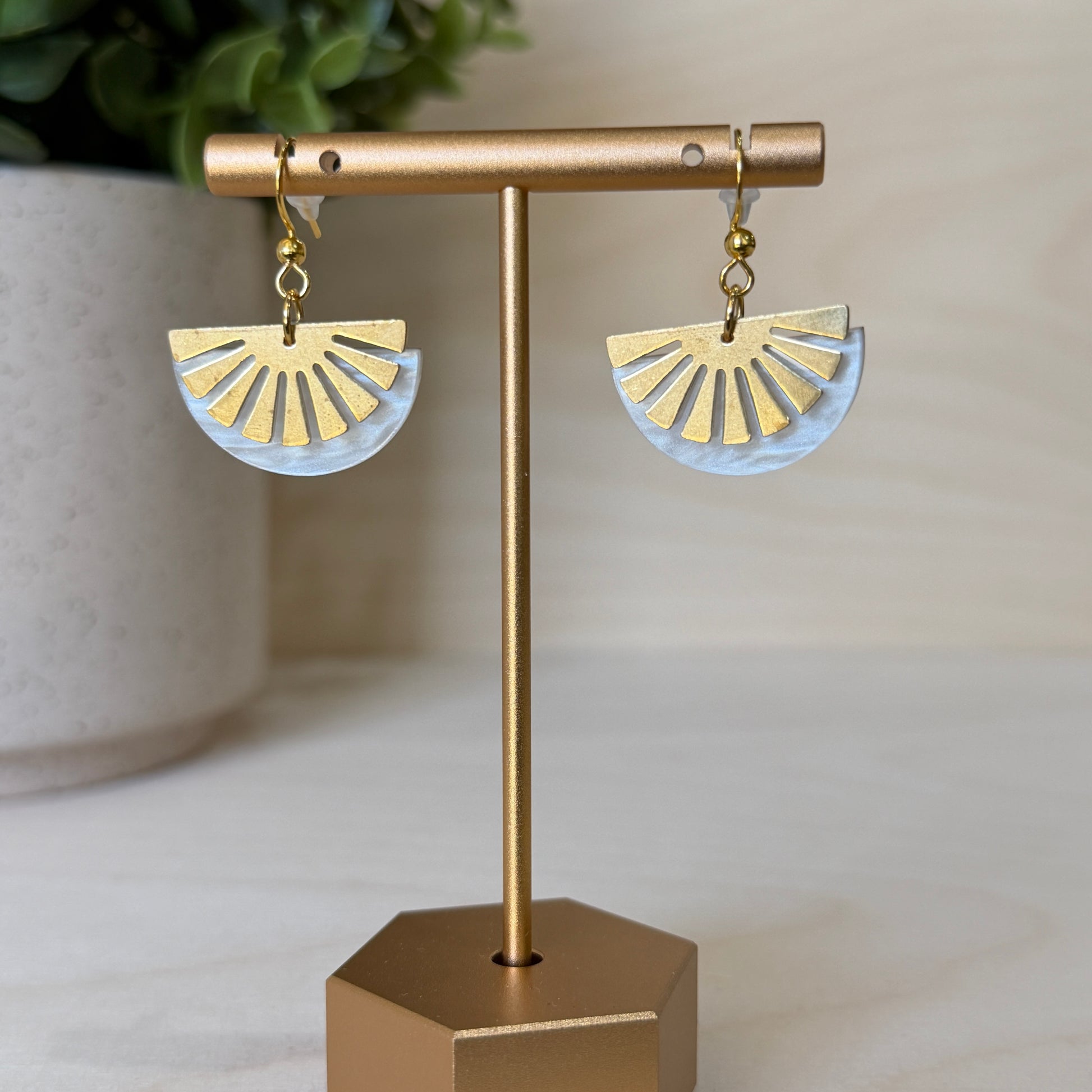 Gold and white half-circle earrings on a gold stand with a plant in the background.