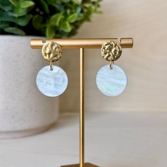 Gold earrings with round, light-colored discs on a gold stand with a blurred plant in the background.