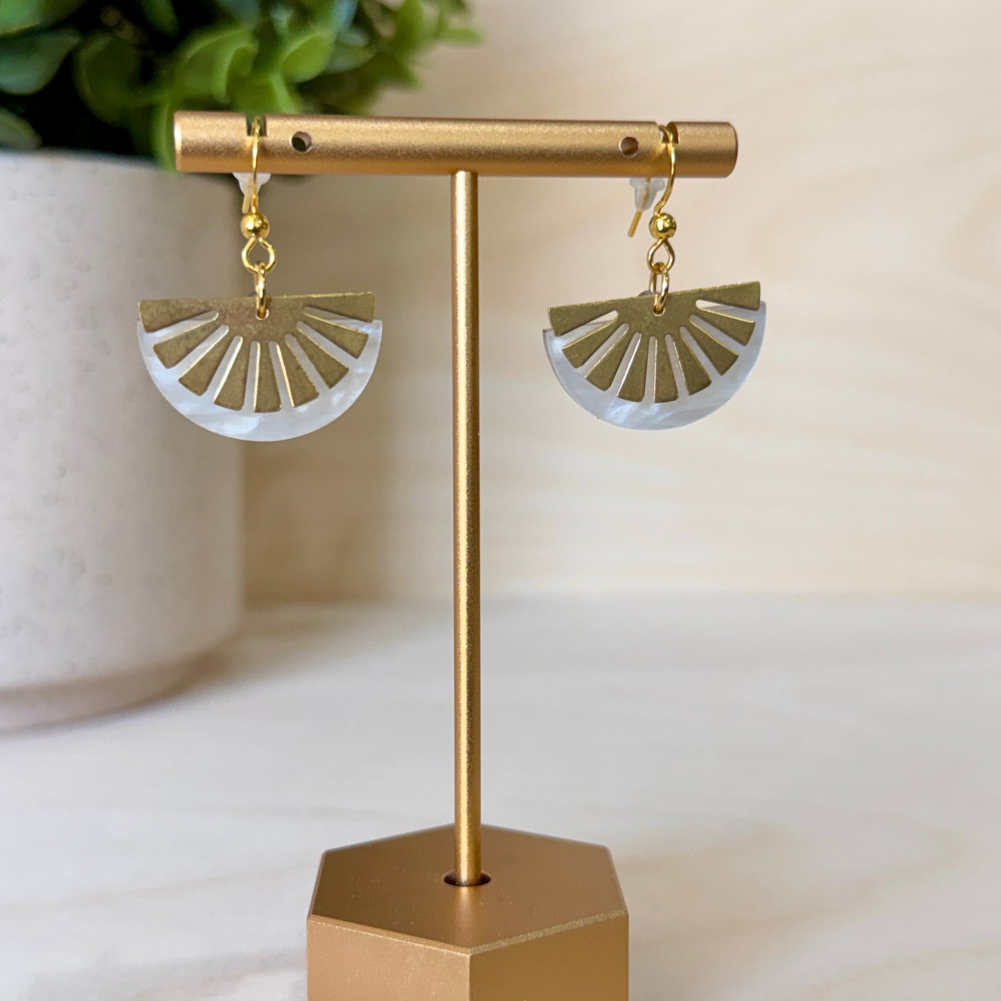 Gold and white earrings on a gold stand with a blurred plant in the background