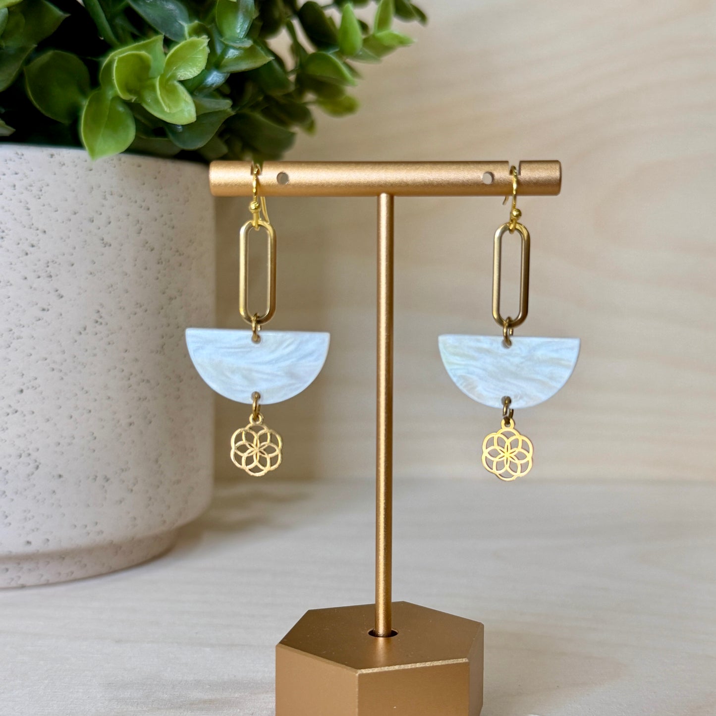 Gold earrings with white semi-circle stones on a gold stand next to a plant.