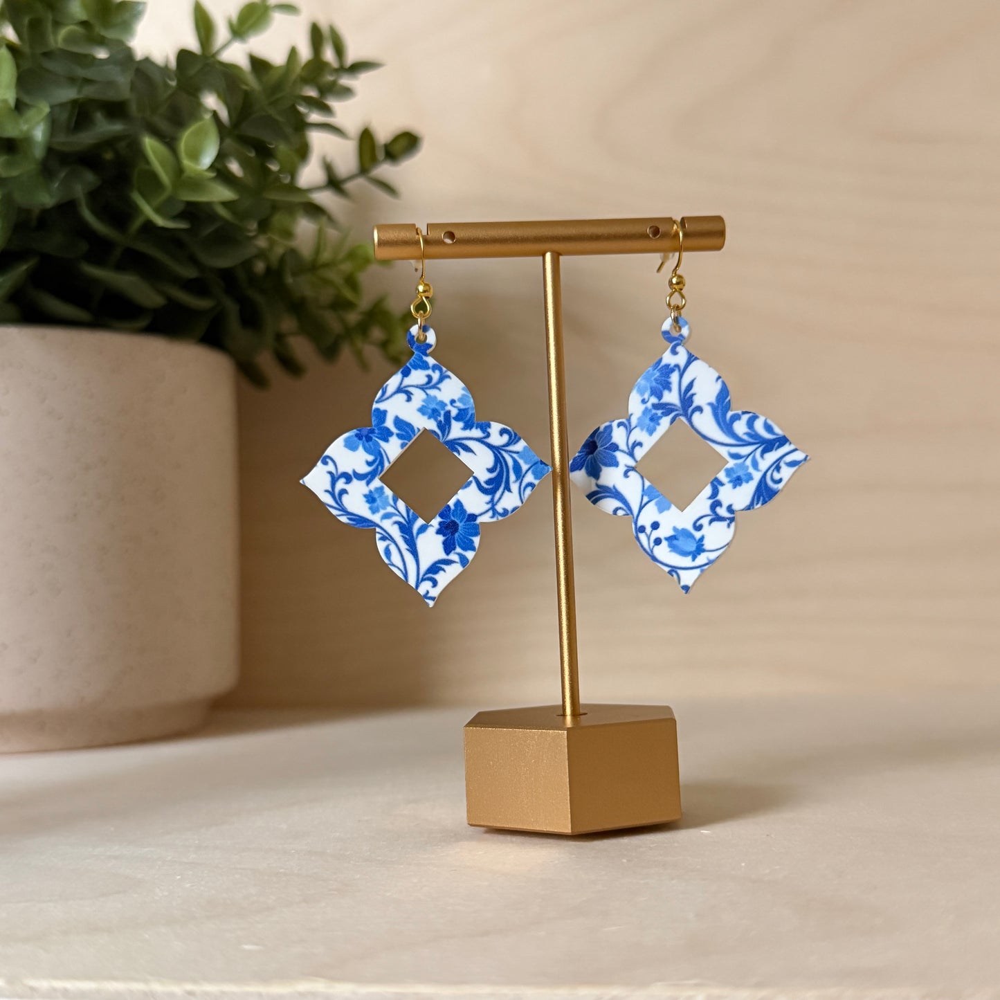 Blue and white patterned earrings on a gold stand with a plant in the background