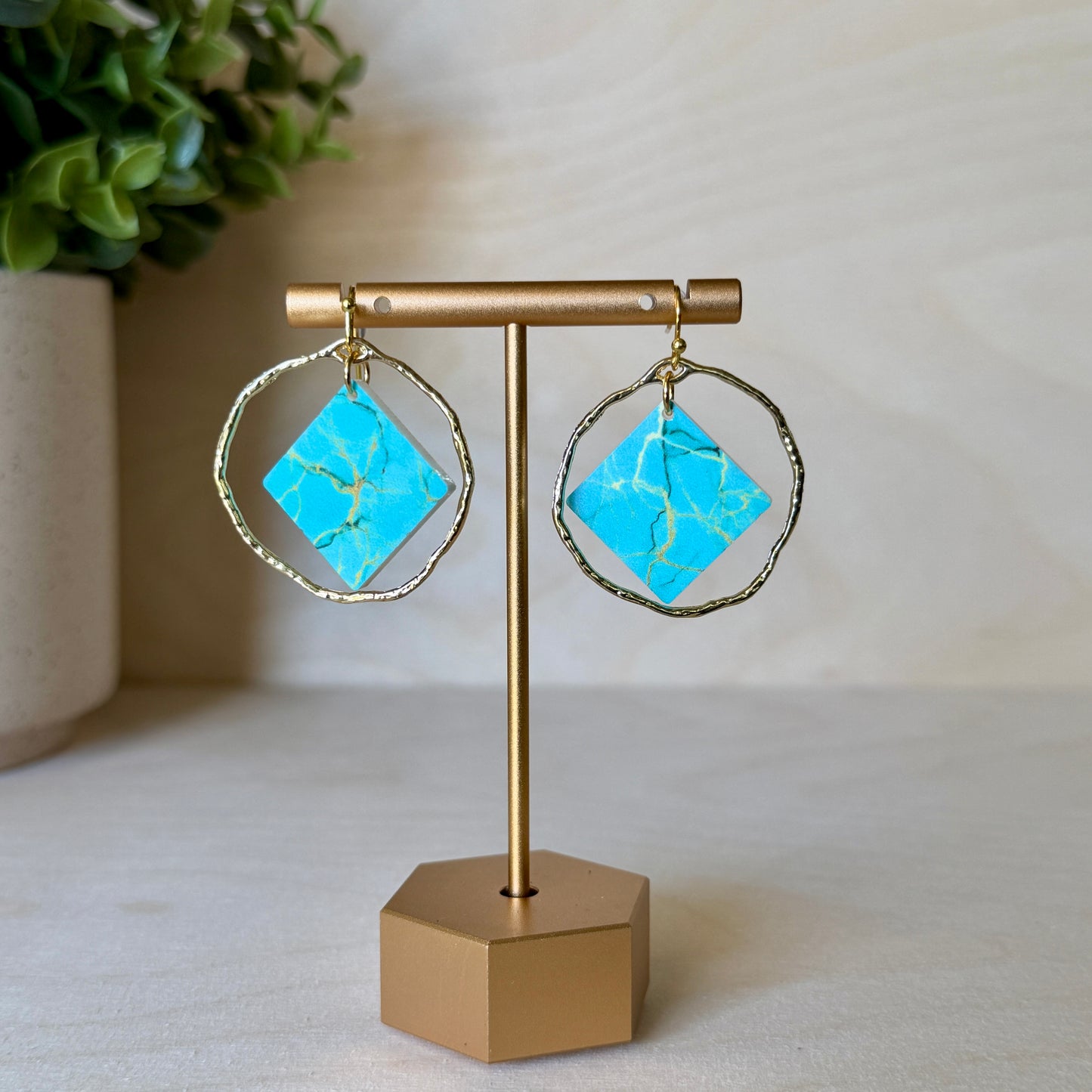 Turquoise square earrings with gold accents on a gold stand against a neutral background