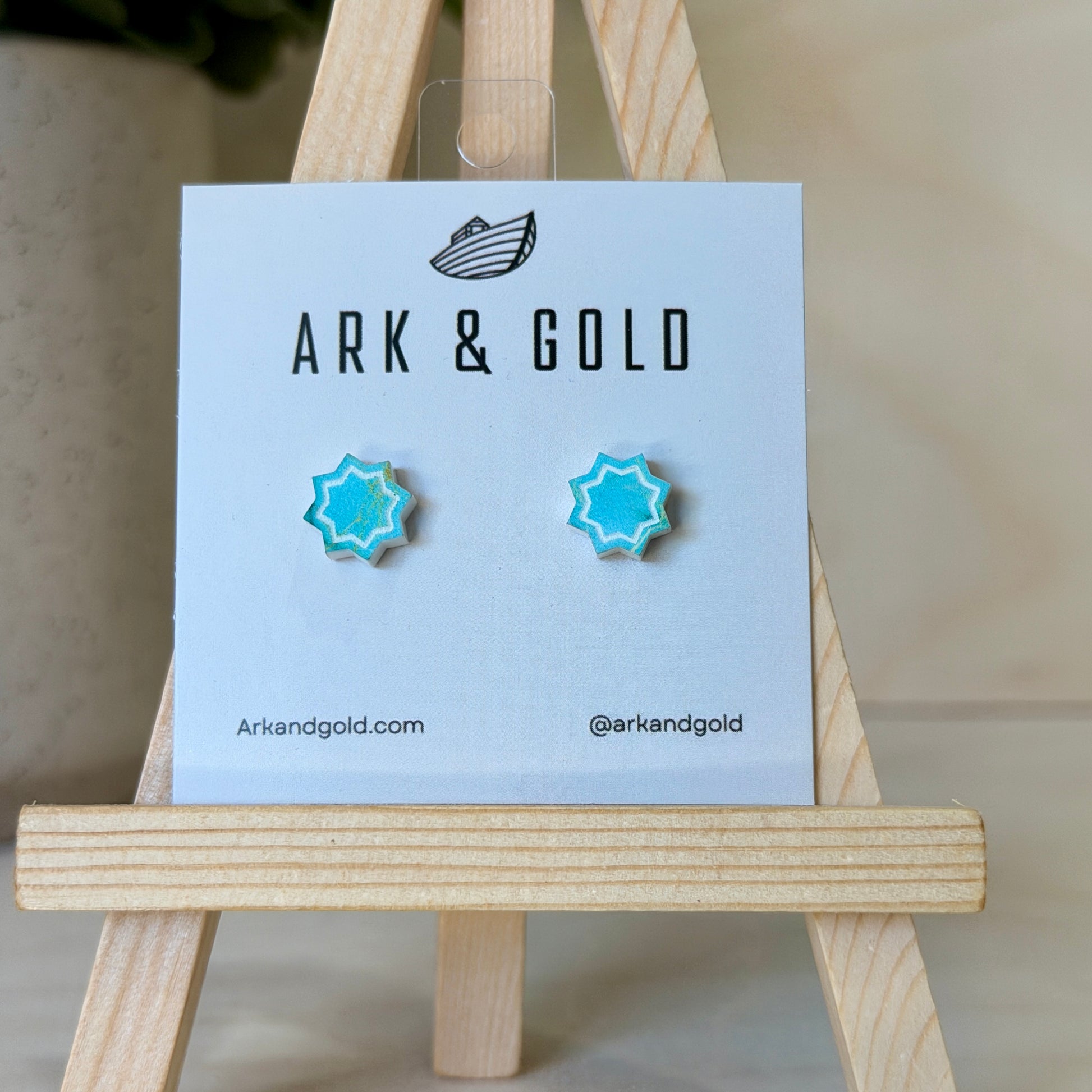 Blue star-shaped earrings on a 'ARK & GOLD' branded card with a wooden stand.