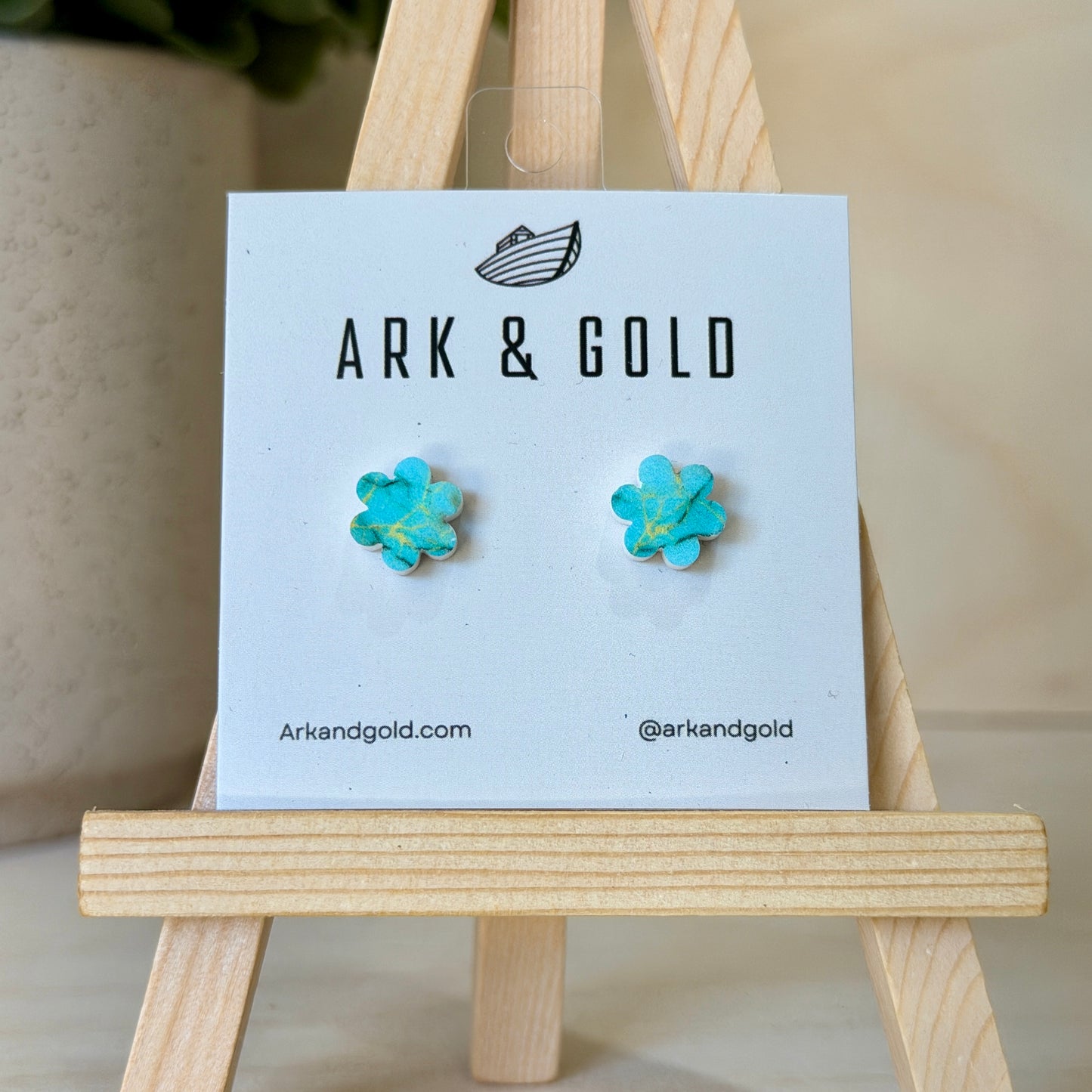 Turquoise flower-shaped earrings on a 'ARK & GOLD' branded card with a wooden stand.