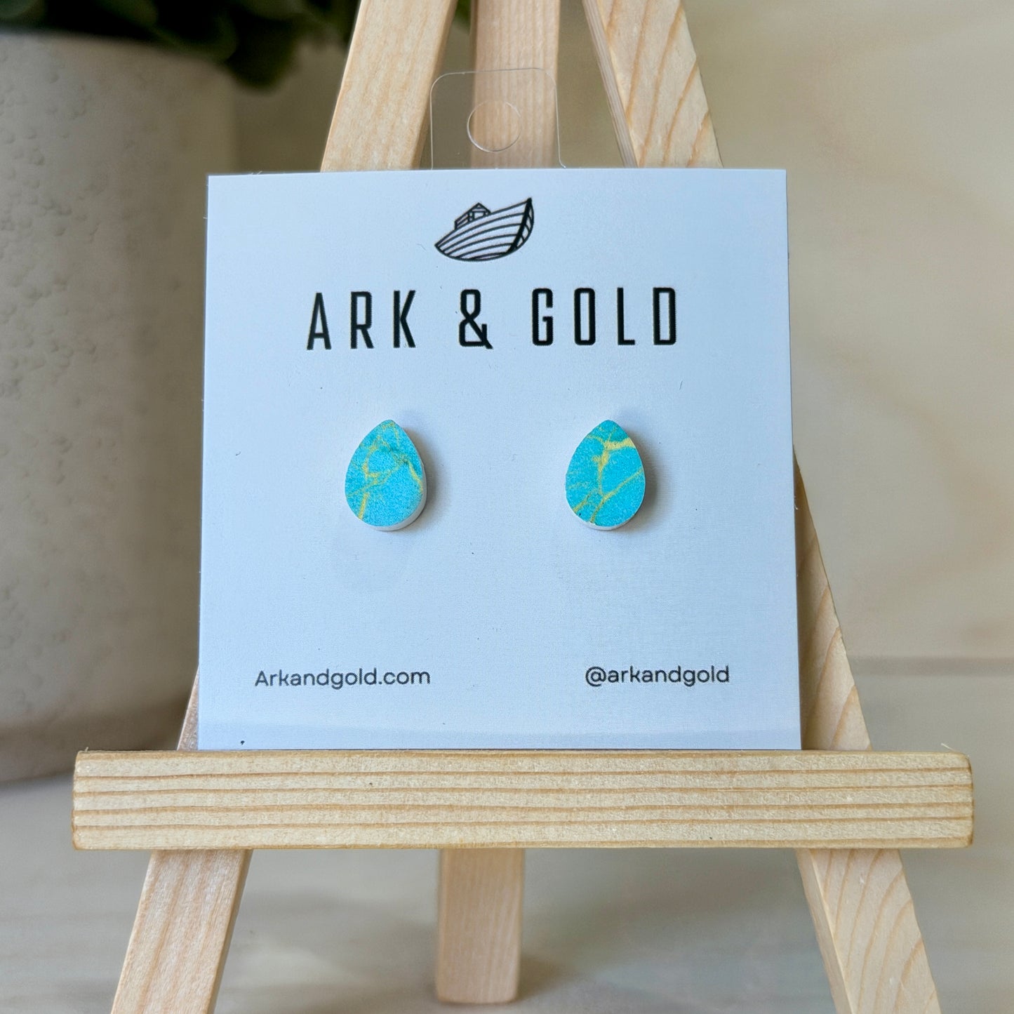 Blue earrings on a 'ARK & GOLD' branded card with a wooden stand.