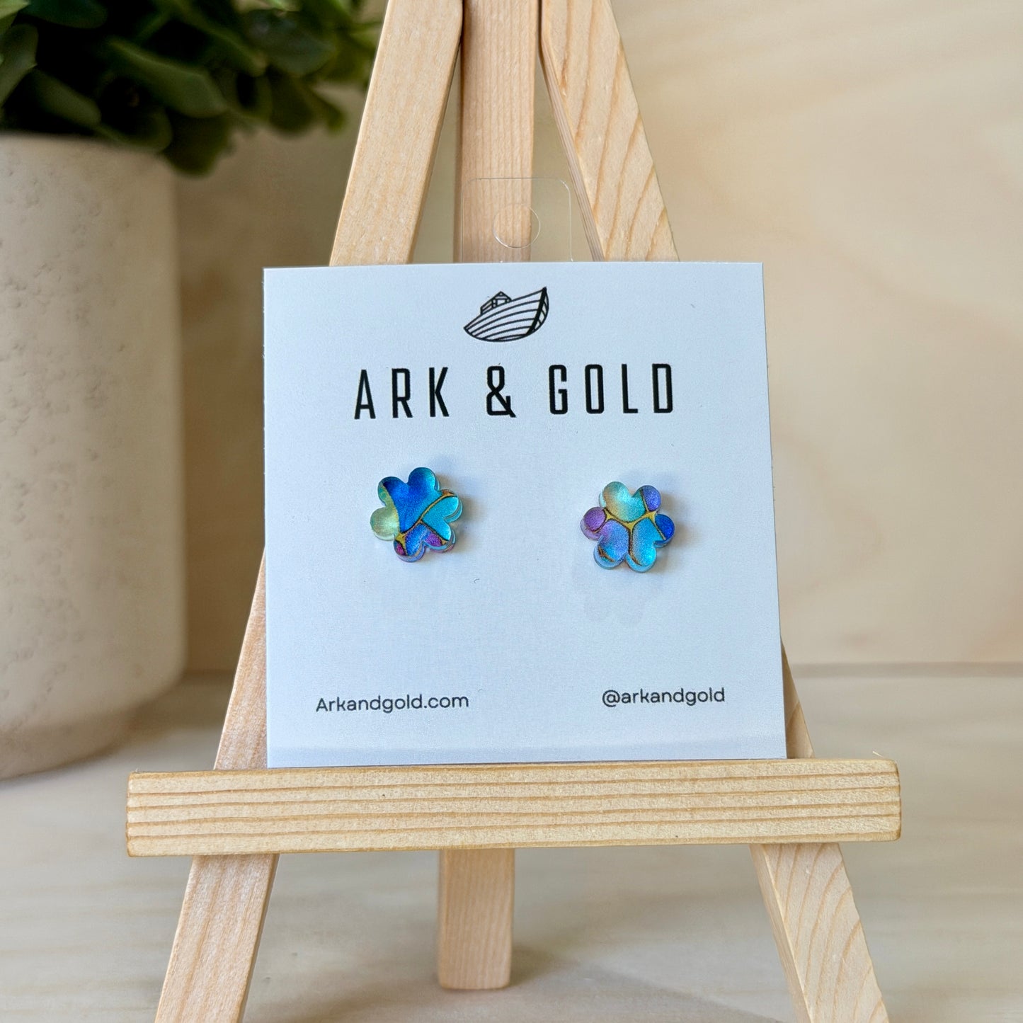 Colorful flower-shaped earrings on a 'ARK & GOLD' branded card with a wooden stand.