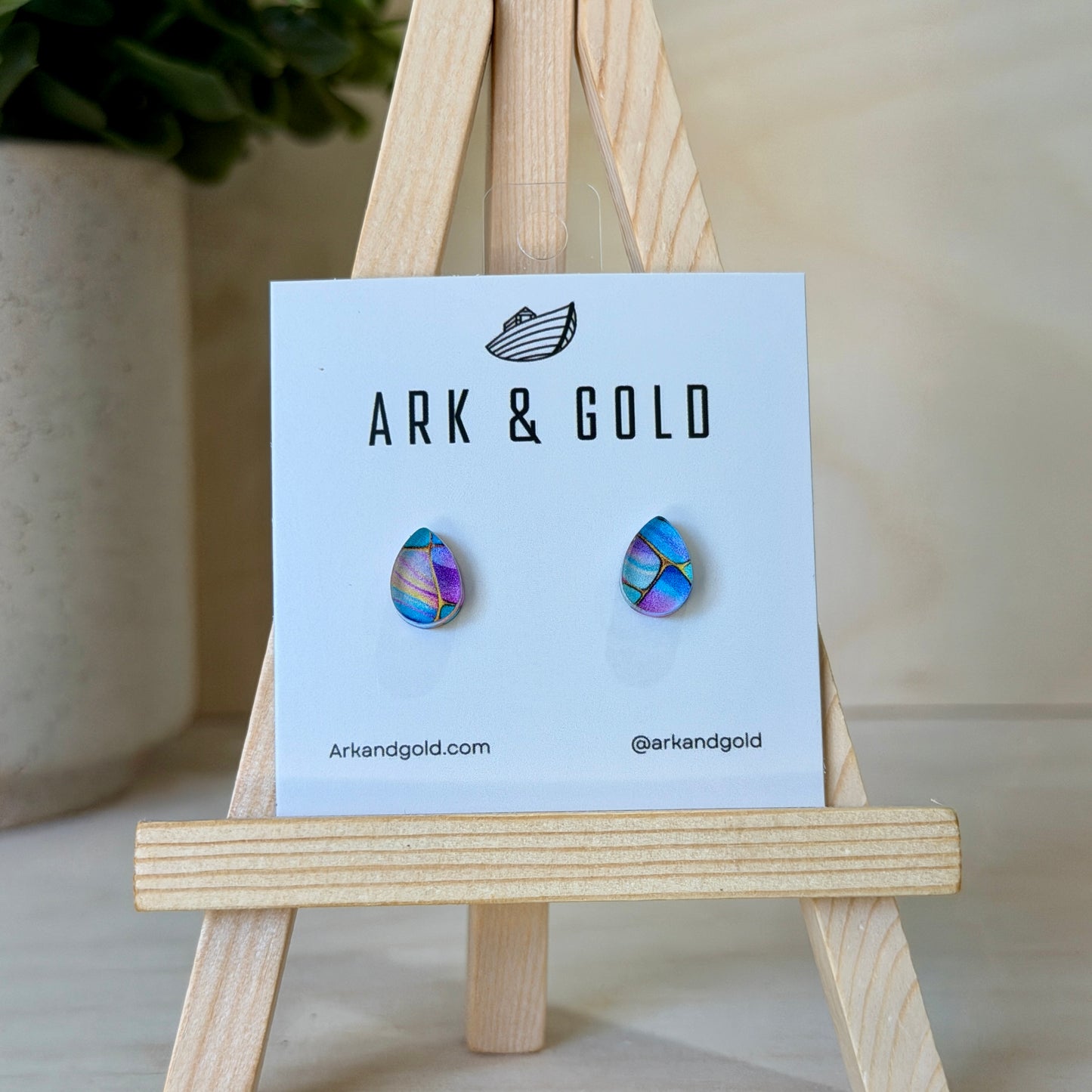Colorful earrings on a 'ARK & GOLD' branded card with a wooden stand.