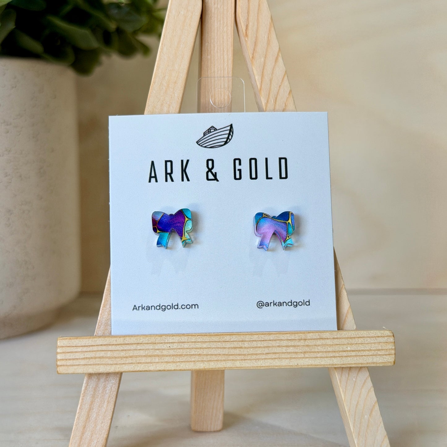 Colorful bow-shaped earrings on a 'ARK & GOLD' branded card with a wooden stand.