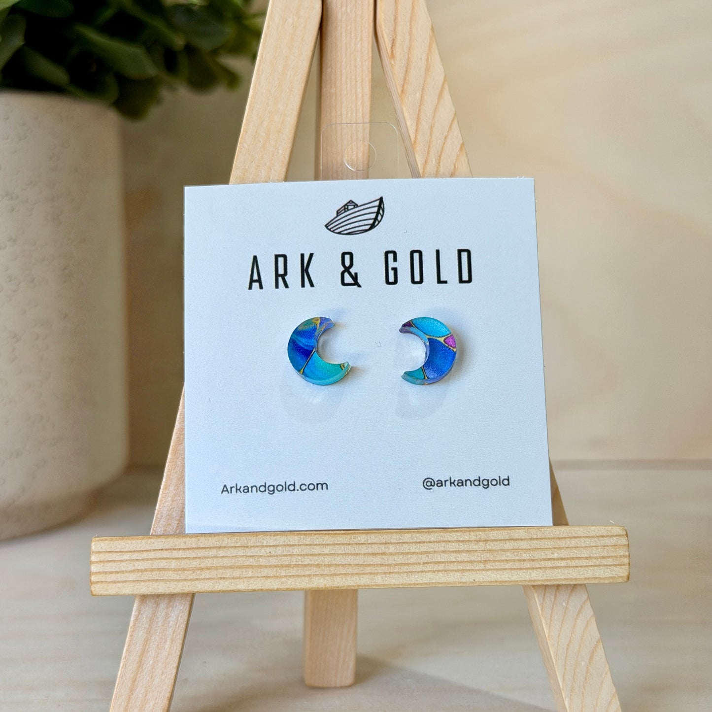 Colorful crescent moon earrings on a 'ARK & GOLD' branded card with a wooden stand.