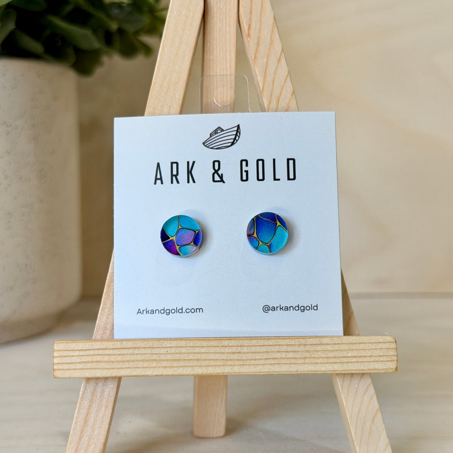Pair of colorful earrings on a 'ARK & GOLD' branded card with a wooden stand.